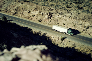 Elevated daytime shot of a green semi‐truck hauling a white box trailer along a sparse desert highway with rocky terrain.
