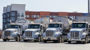 Four polished Peterbilt semi‑trucks with bulk grain trailers parked in an industrial yard.