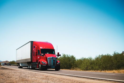 Red Class 8 semi truck hauling a trailer along a rural highway under daylight skies.