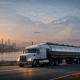 Stainless steel fuel tanker semi-truck driving on a highway at sunrise with a refinery skyline and distant cargo ships on calm water.