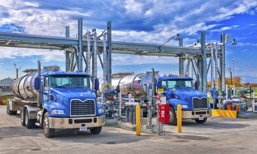 Two tanker trucks positioned at a fuel-loading terminal with overhead piping and transfer equipment.