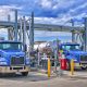 Two tanker trucks positioned at a fuel-loading terminal with overhead piping and transfer equipment.
