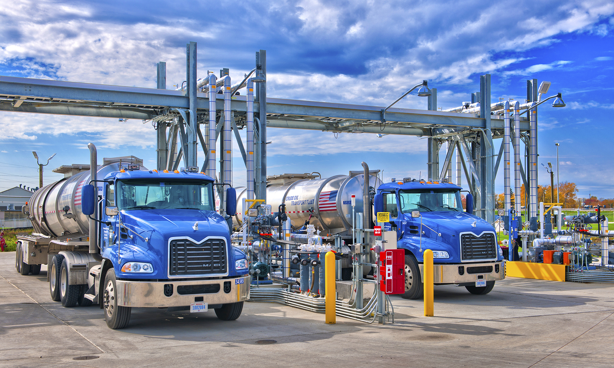 Two tanker trucks positioned at a fuel-loading terminal with overhead piping and transfer equipment.