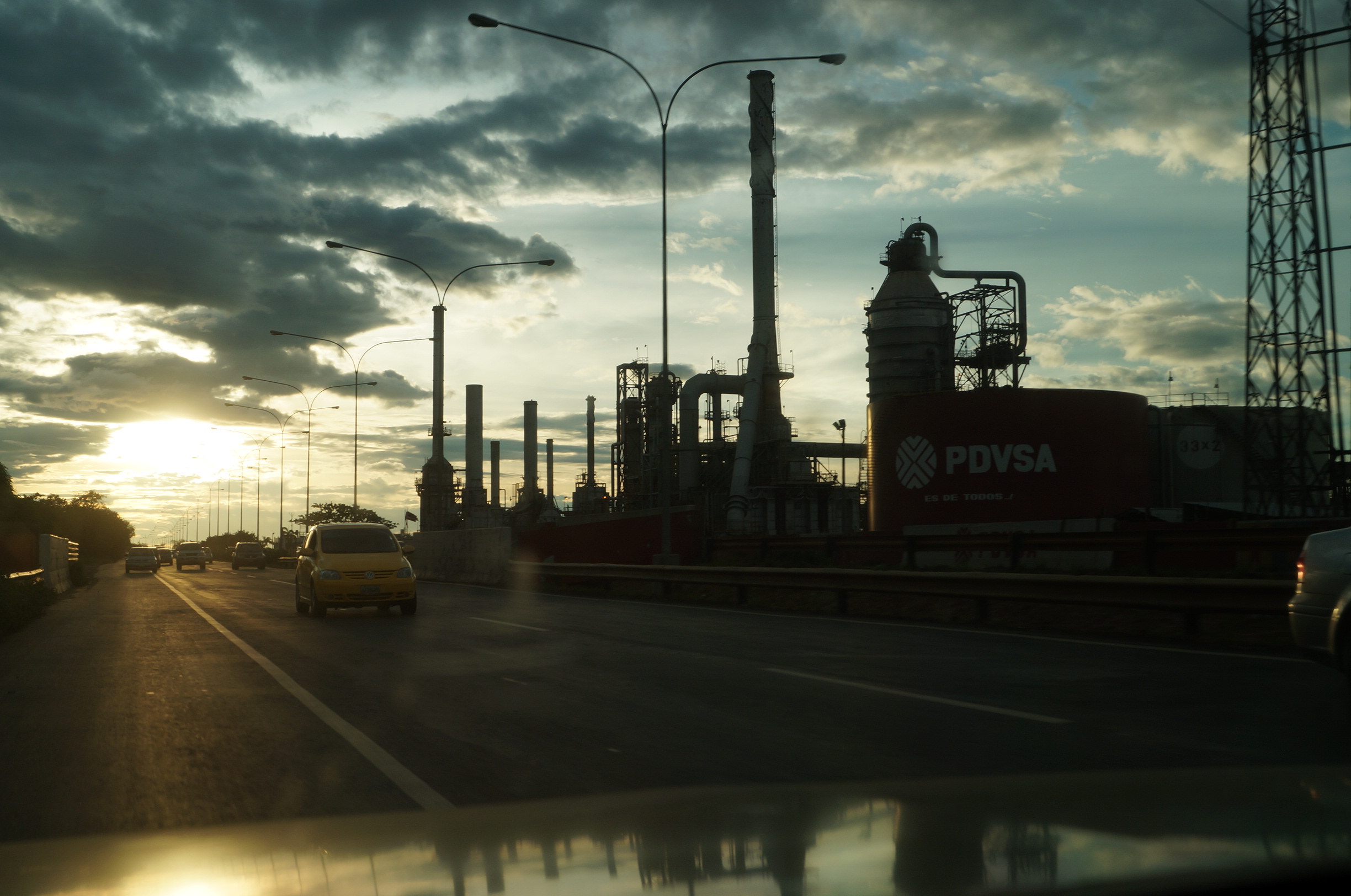 El Palito refinery in Venezuela at sunset, with roadside traffic, industrial towers and a red PDVSA storage tank.