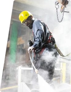 Trimac Adds Houston-Area ISO Tank Depot With 600-Tank Capacity 3 Worker wearing a yellow hard hat, safety glasses and harness uses a spray lance amid mist on an elevated platform.